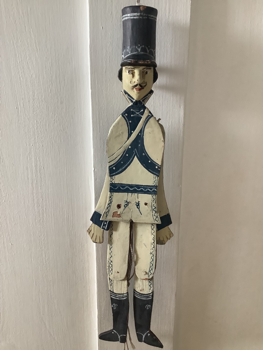 A double-sided flat carved wooden soldier puppet featuring a painted blue jacket, red trousers, black boots, tall black shako hat, and central hanging string. View II