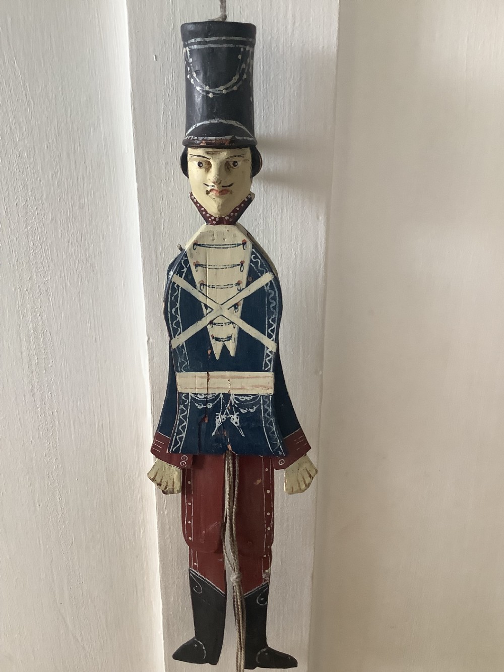 A double-sided flat carved wooden soldier puppet featuring a painted blue jacket, red trousers, black boots, tall black shako hat, and central hanging string.