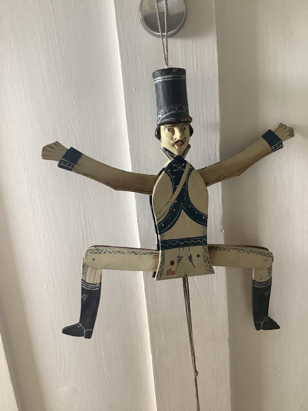 A double-sided flat carved wooden soldier puppet featuring a painted blue jacket, red trousers, black boots, tall black shako hat, and central hanging string. View III