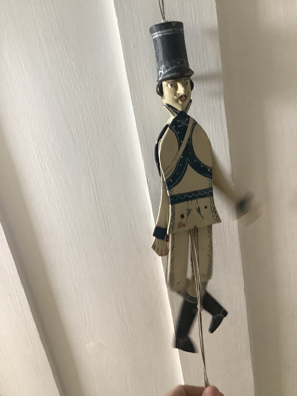 A double-sided flat carved wooden soldier puppet featuring a painted blue jacket, red trousers, black boots, tall black shako hat, and central hanging string. View IV