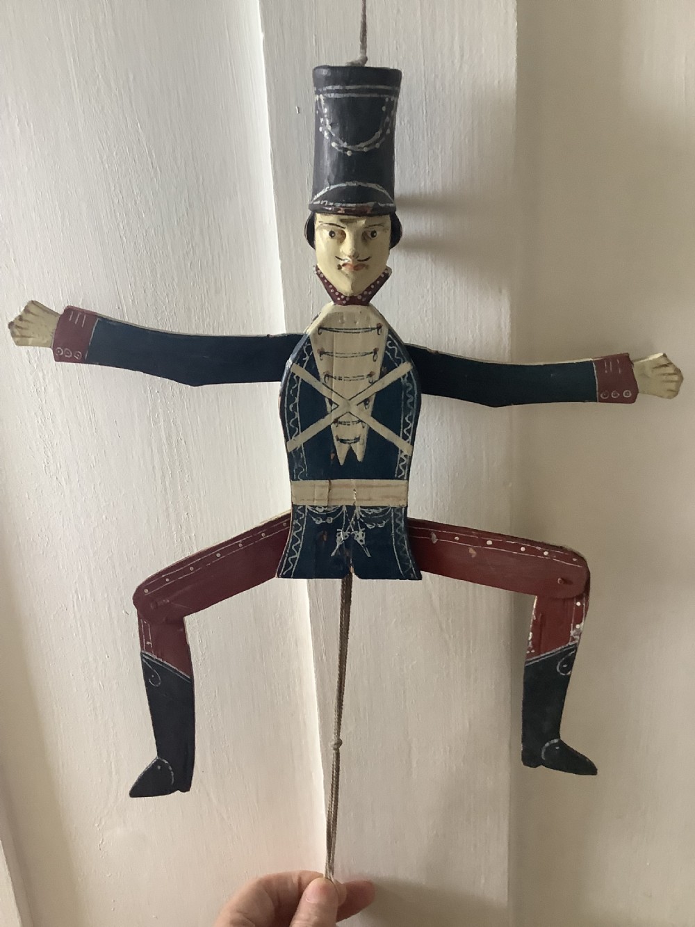 A double-sided flat carved wooden soldier puppet featuring a painted blue jacket, red trousers, black boots, tall black shako hat, and central hanging string. View V