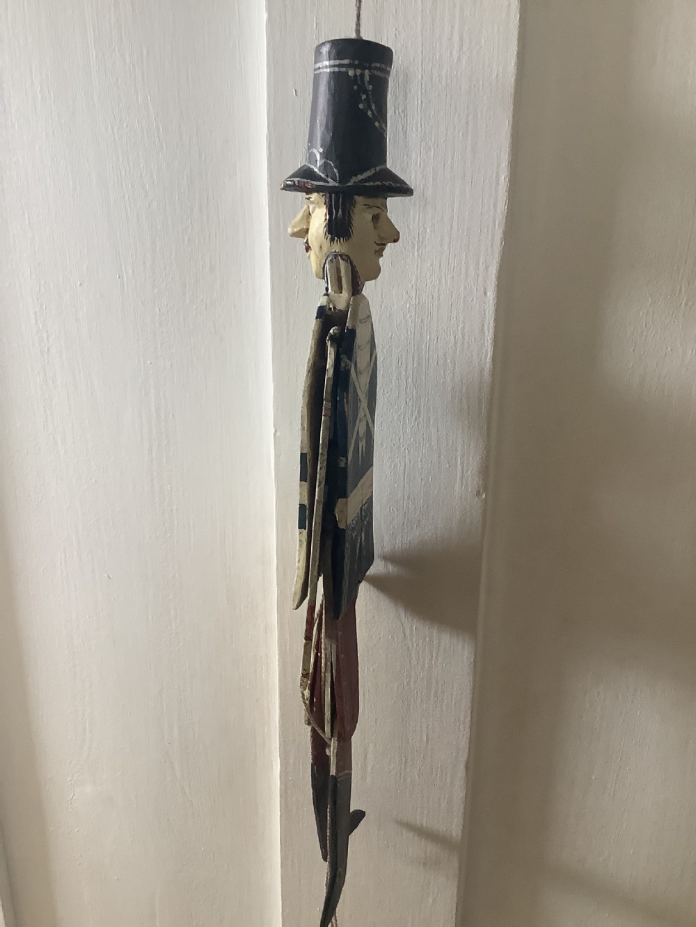A double-sided flat carved wooden soldier puppet featuring a painted blue jacket, red trousers, black boots, tall black shako hat, and central hanging string. View VI
