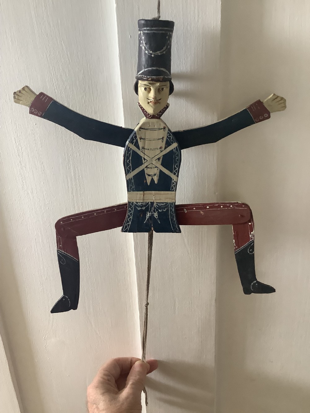 A double-sided flat carved wooden soldier puppet featuring a painted blue jacket, red trousers, black boots, tall black shako hat, and central hanging string. View VII