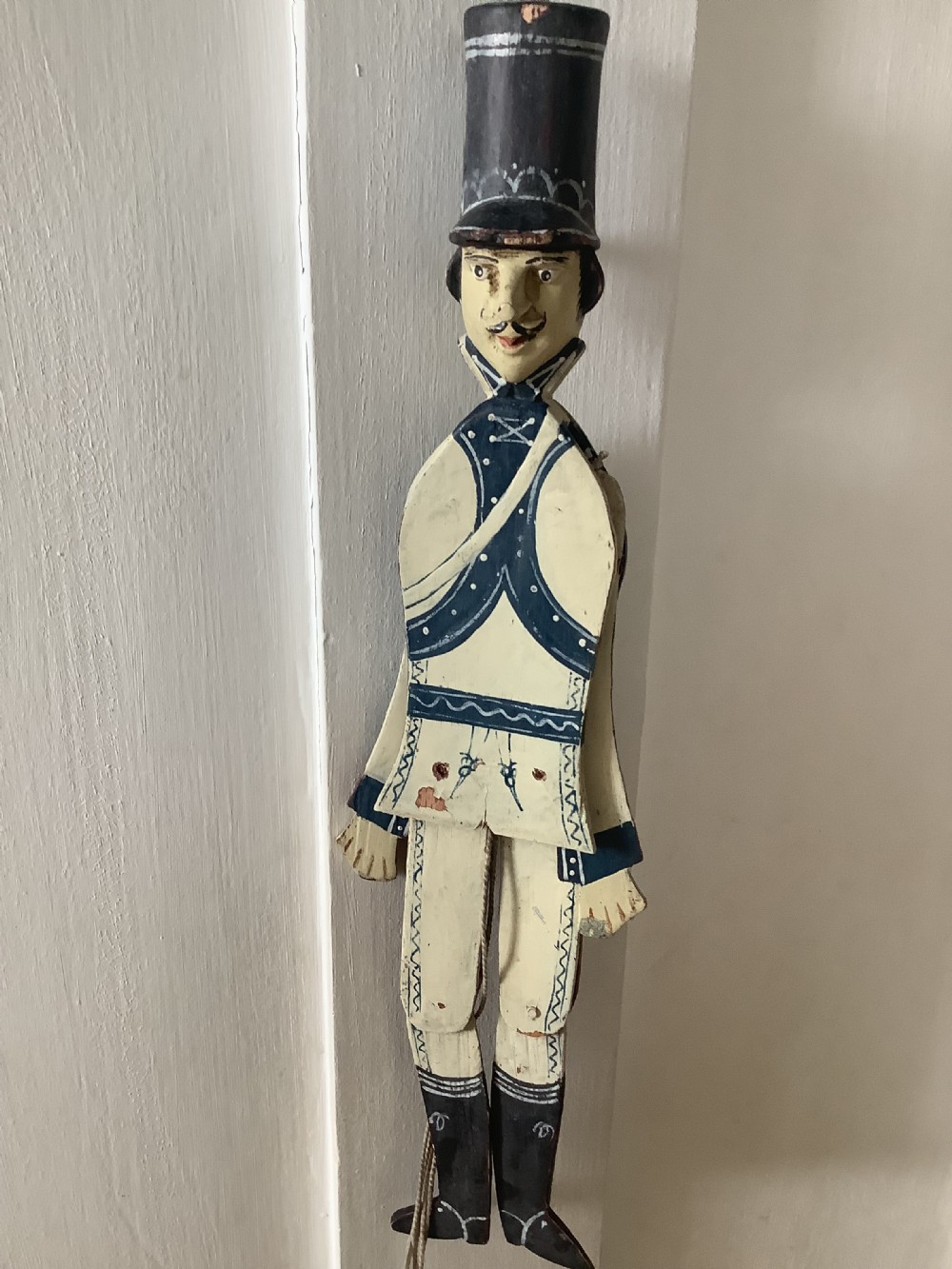 A double-sided flat carved wooden soldier puppet featuring a painted blue jacket, red trousers, black boots, tall black shako hat, and central hanging string. View X