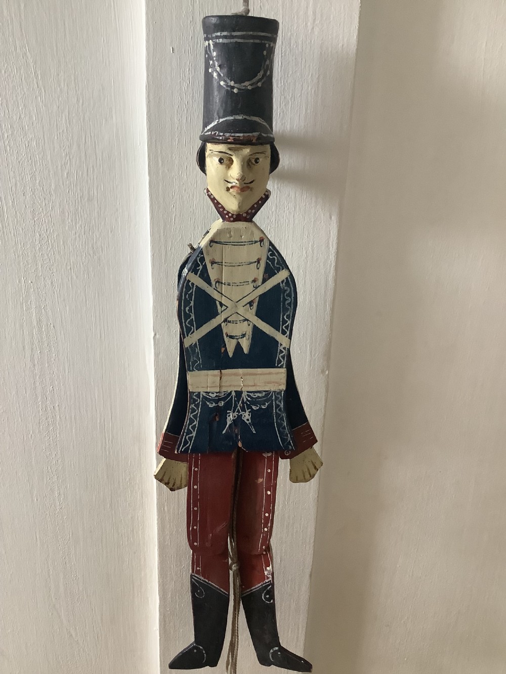 A double-sided flat carved wooden soldier puppet featuring a painted blue jacket, red trousers, black boots, tall black shako hat, and central hanging string. View XI