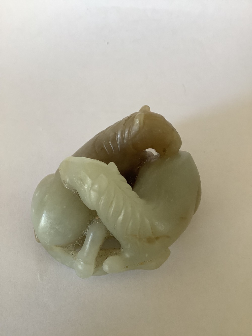 A small 19th-century Chinese hand-carved jade sculpture depicting two intertwined recumbent horses in pale celadon green and grey hardstone.