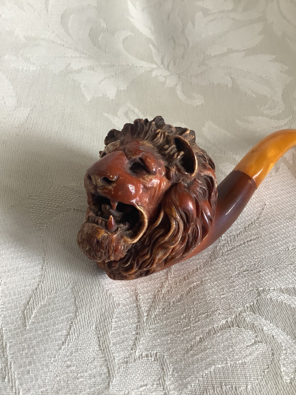 A hand-carved meerschaum smoking pipe featuring a detailed lion head bowl with an open mouth and a translucent amber-coloured curved stem.