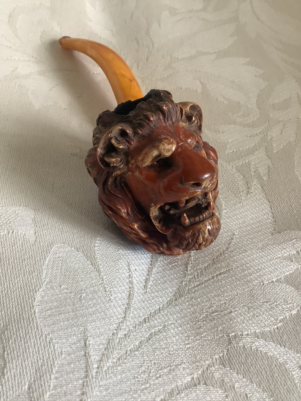 A hand-carved meerschaum smoking pipe featuring a detailed lion head bowl with an open mouth and a translucent amber-coloured curved stem. View VI