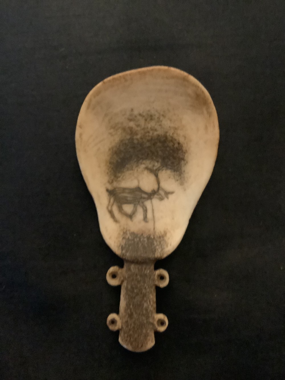 A hand-carved bone or antler spoon featuring an etched drawing of a caribou on the shallow cream-coloured bowl and a textured handle with four side loops.
