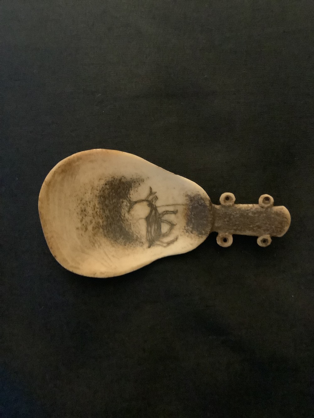 A hand-carved bone or antler spoon featuring an etched drawing of a caribou on the shallow cream-coloured bowl and a textured handle with four side loops. View III