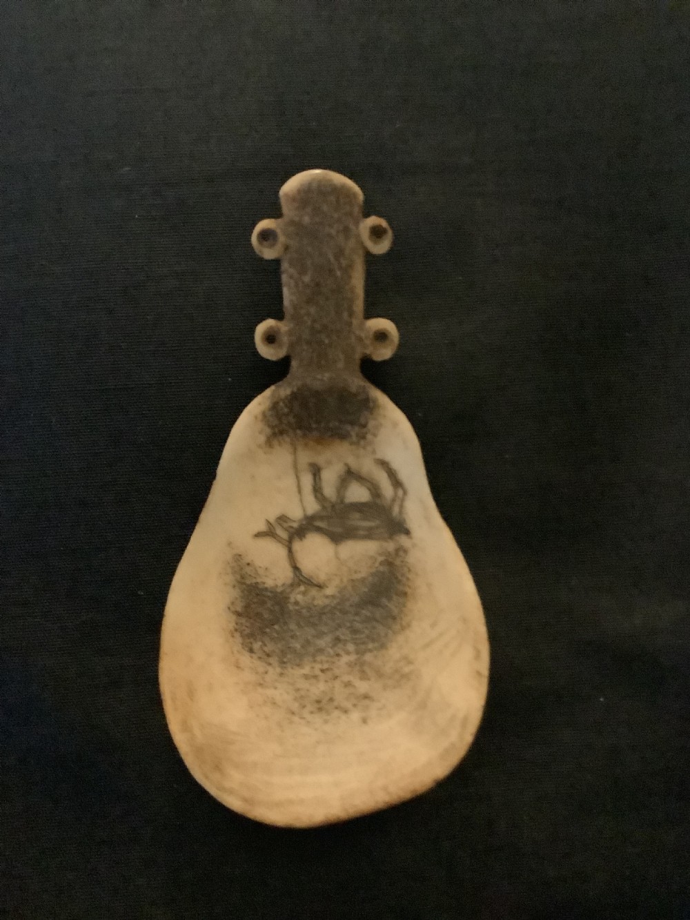 A hand-carved bone or antler spoon featuring an etched drawing of a caribou on the shallow cream-coloured bowl and a textured handle with four side loops. View IV