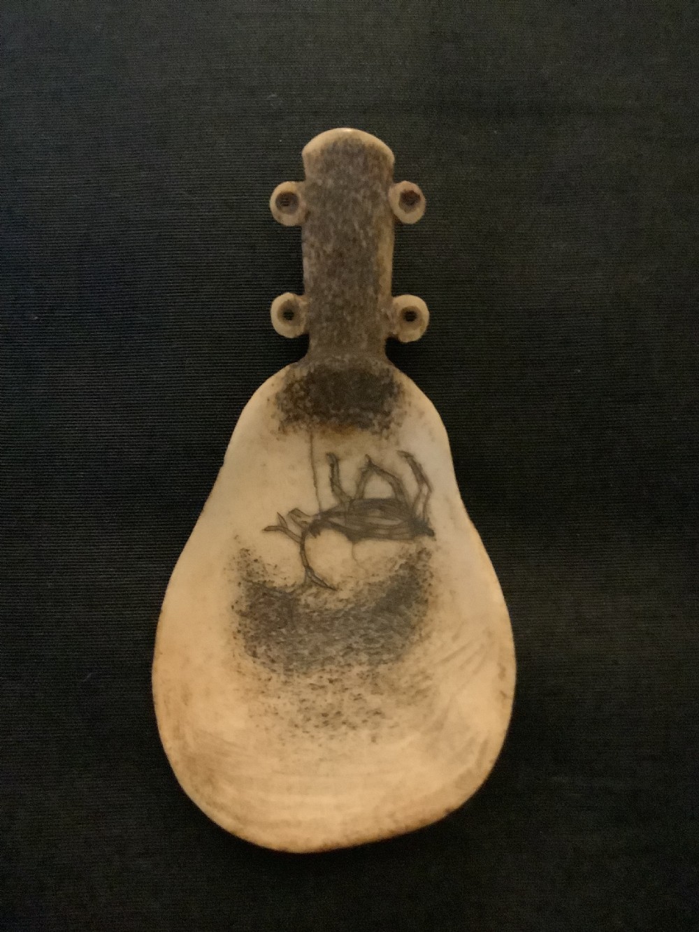 A hand-carved bone or antler spoon featuring an etched drawing of a caribou on the shallow cream-coloured bowl and a textured handle with four side loops. View VI