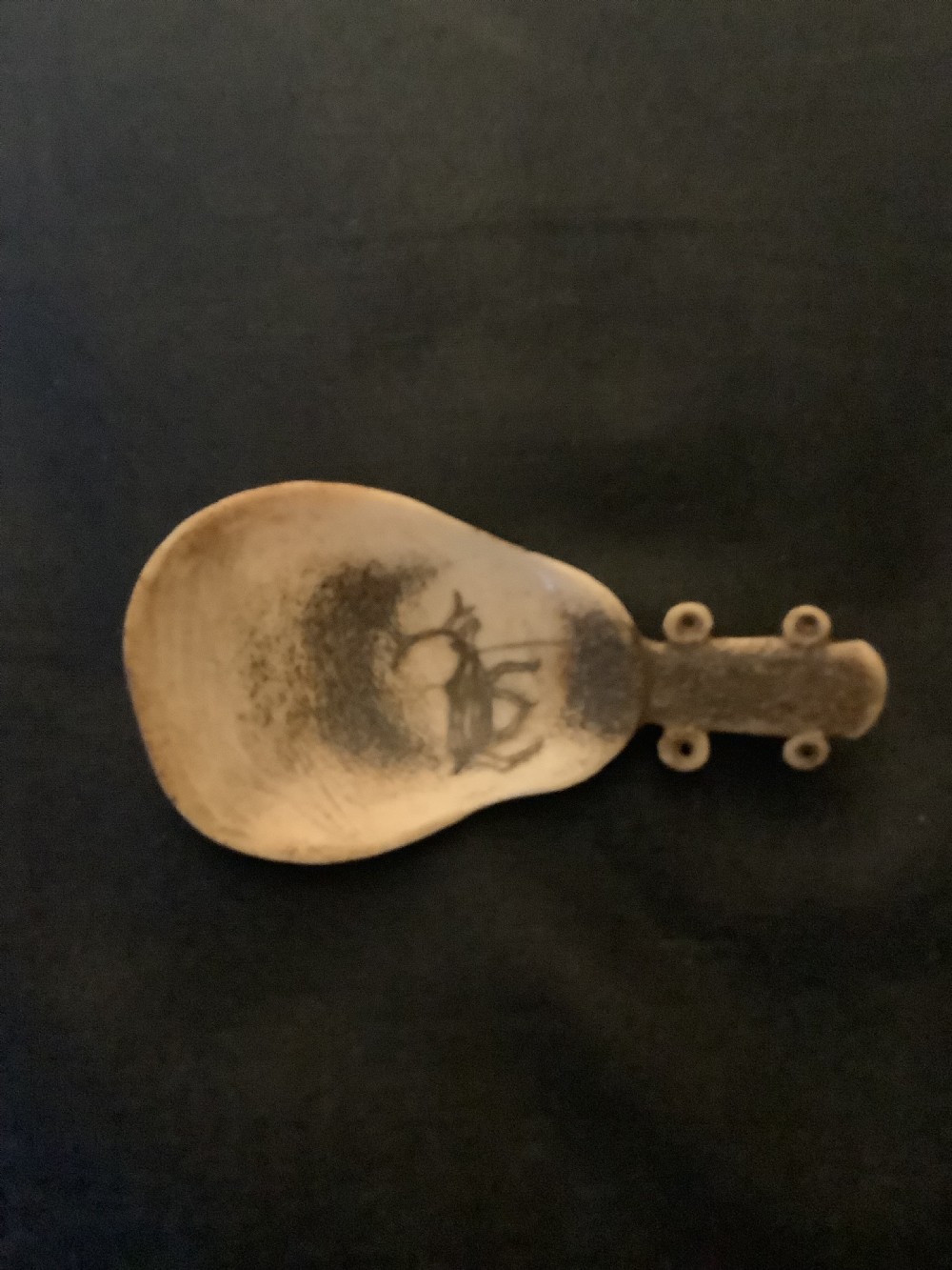 A hand-carved bone or antler spoon featuring an etched drawing of a caribou on the shallow cream-coloured bowl and a textured handle with four side loops. View VIII
