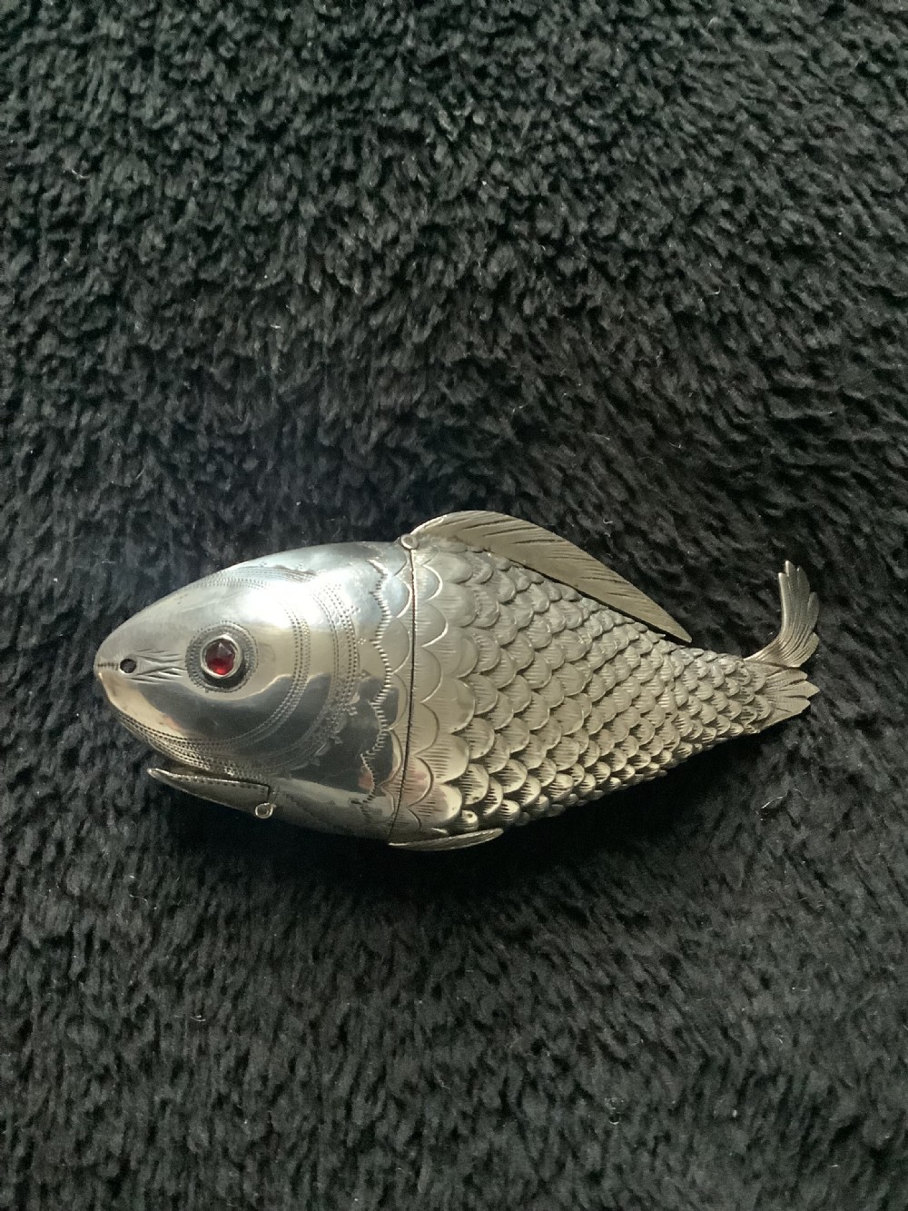 A silver fish-shaped box featuring engraved scales, an articulated body, a hinged head opening, and a red glass eye. View II