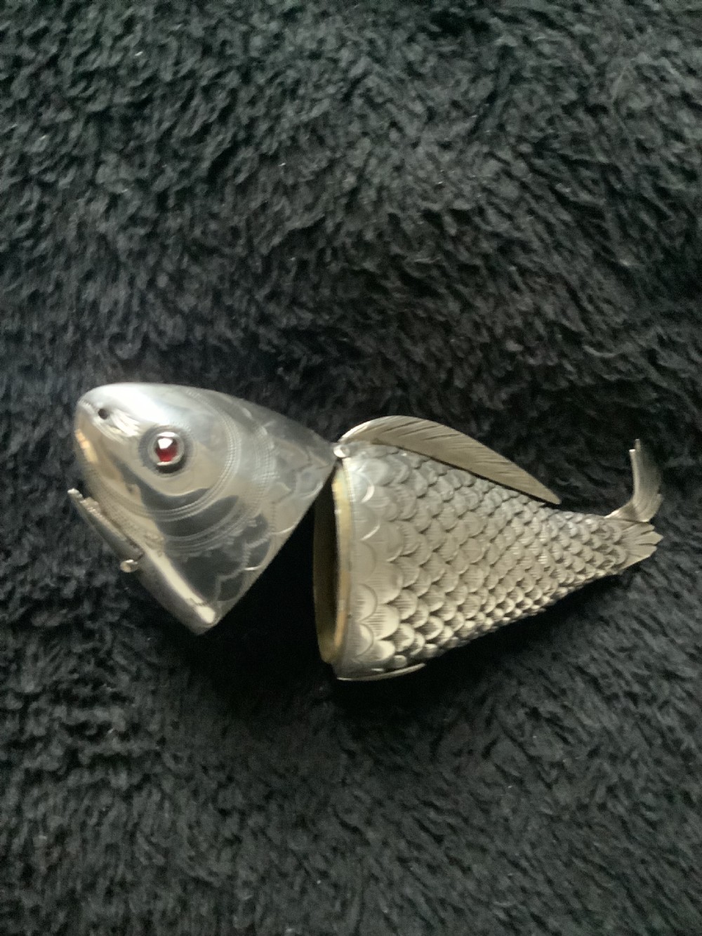 A silver fish-shaped box featuring engraved scales, an articulated body, a hinged head opening, and a red glass eye. View III