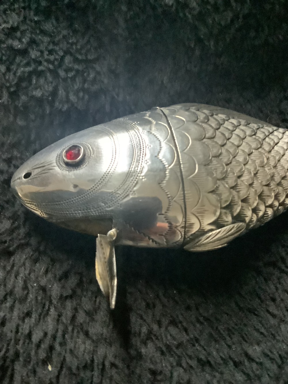 A silver fish-shaped box featuring engraved scales, an articulated body, a hinged head opening, and a red glass eye. View IV