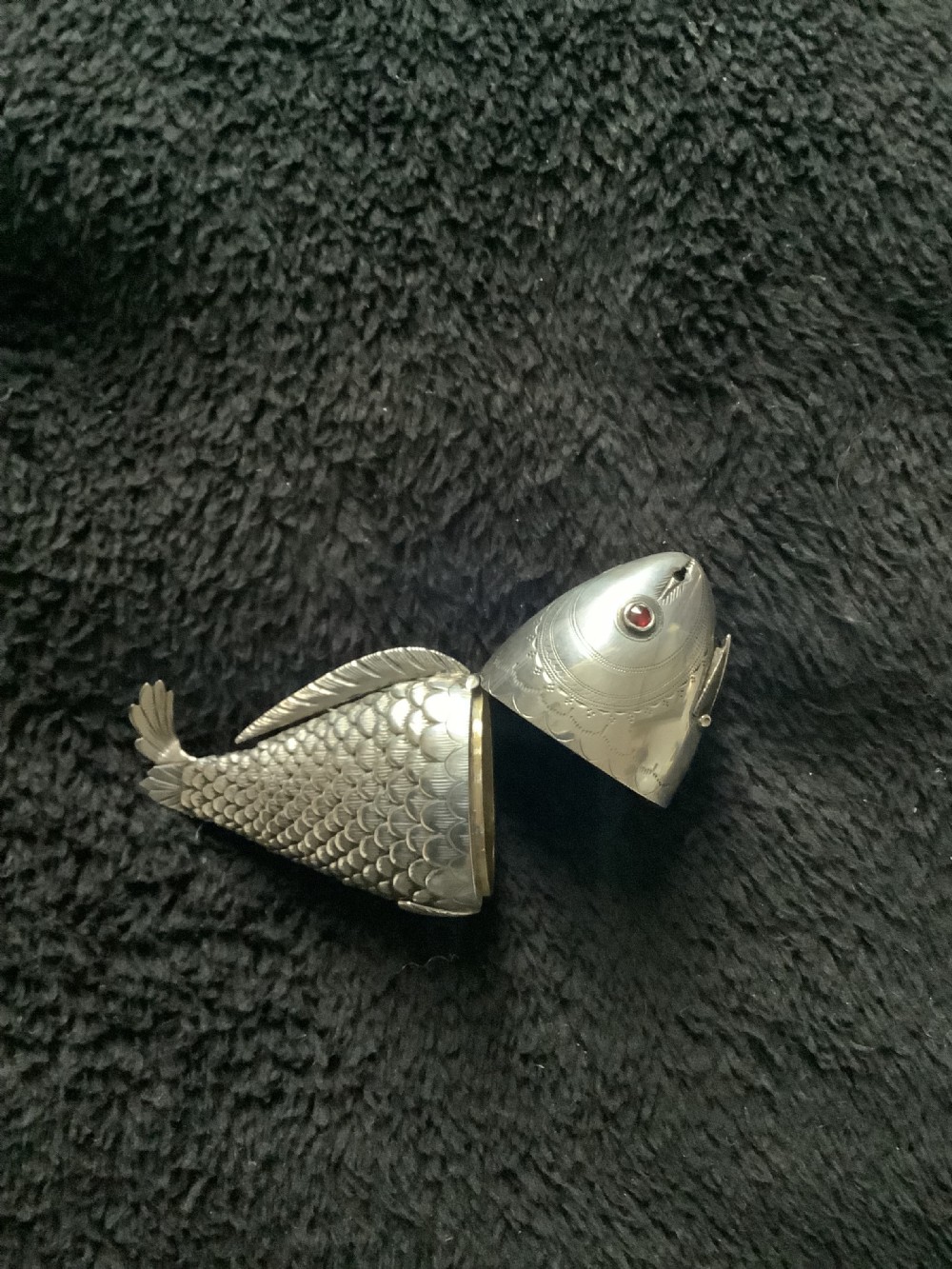 A silver fish-shaped box featuring engraved scales, an articulated body, a hinged head opening, and a red glass eye. View V