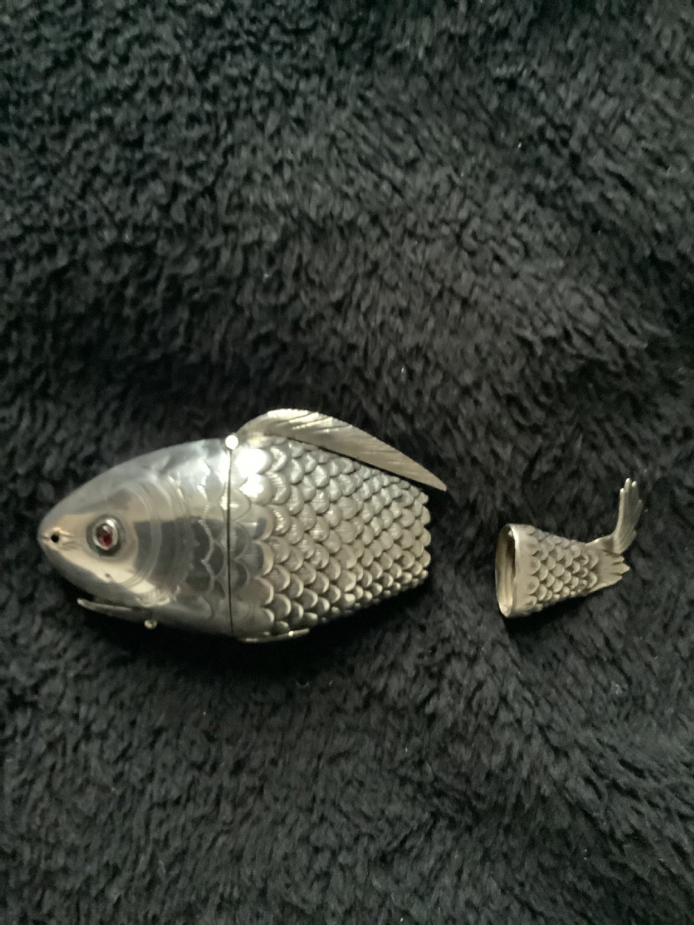 A silver fish-shaped box featuring engraved scales, an articulated body, a hinged head opening, and a red glass eye. View VI