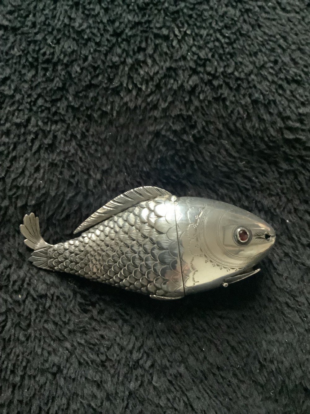 A silver fish-shaped box featuring engraved scales, an articulated body, a hinged head opening, and a red glass eye. View VII