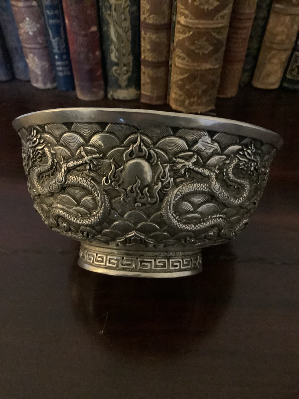 A circular silver-coloured metal bowl decorated with high-relief Chinese dragons chasing a flaming pearl above a key fret foot rim.