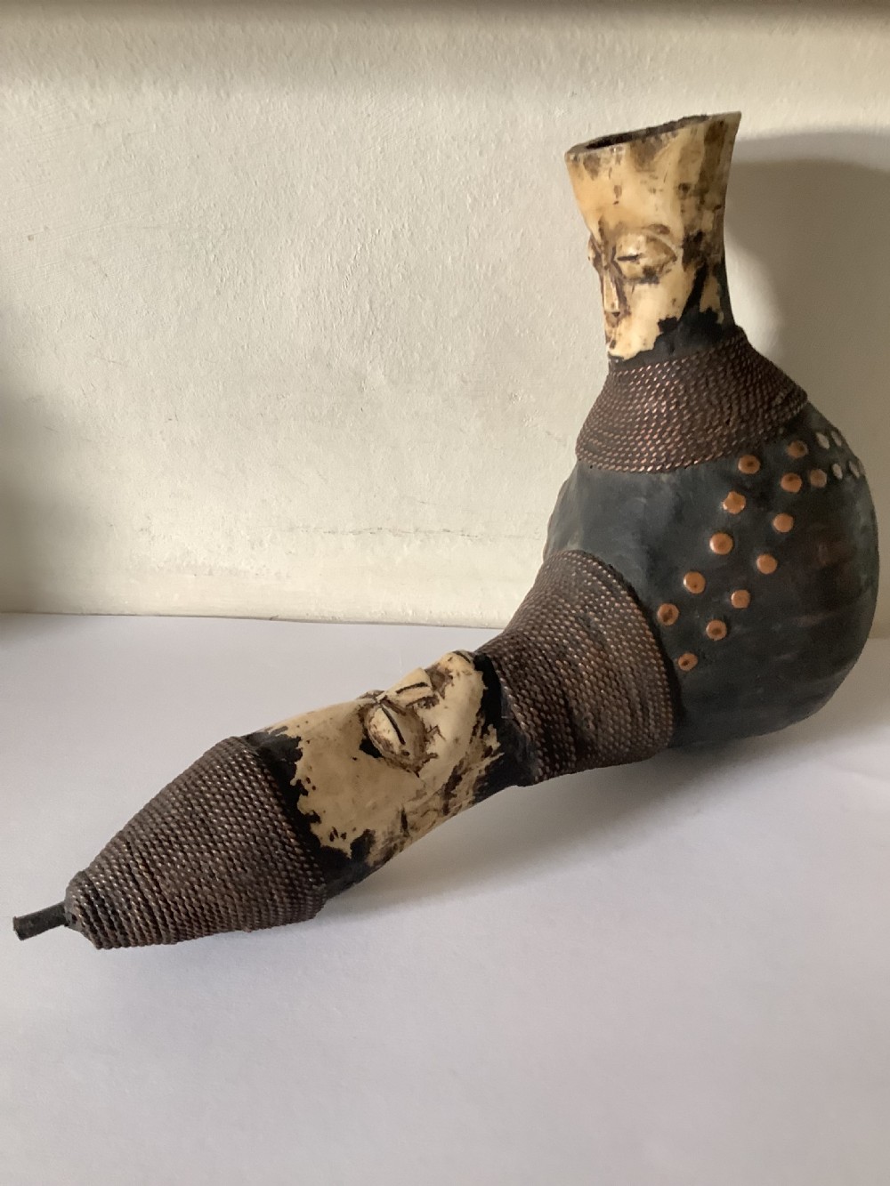 A hand-carved African smoking pipe featuring a dark rounded body with metal studs, woven wire binding, and stylised carved bone faces on the bowl and stem. View V