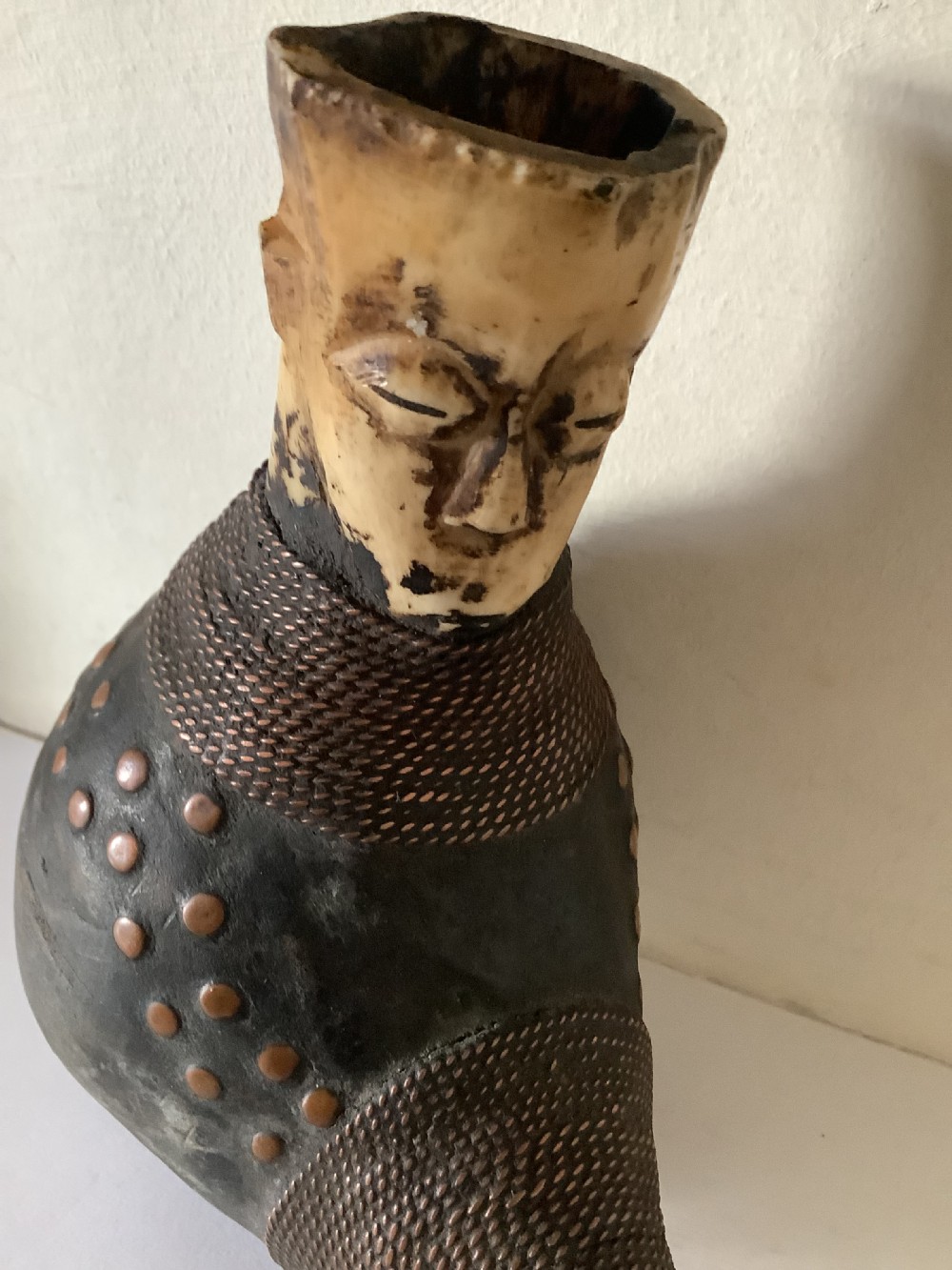 A hand-carved African smoking pipe featuring a dark rounded body with metal studs, woven wire binding, and stylised carved bone faces on the bowl and stem. View II