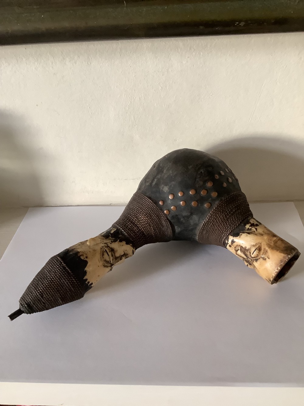 A hand-carved African smoking pipe featuring a dark rounded body with metal studs, woven wire binding, and stylised carved bone faces on the bowl and stem. View III
