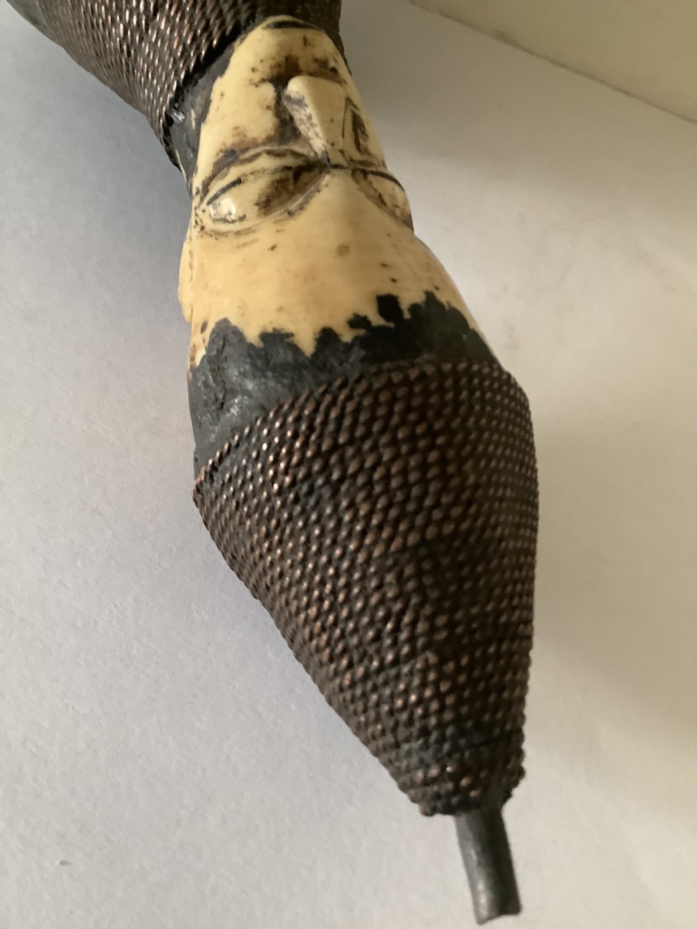 A hand-carved African smoking pipe featuring a dark rounded body with metal studs, woven wire binding, and stylised carved bone faces on the bowl and stem. View IV