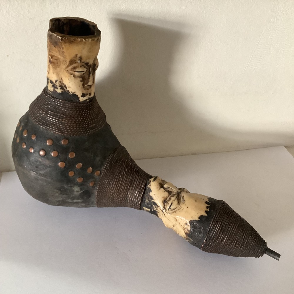 A hand-carved African smoking pipe featuring a dark rounded body with metal studs, woven wire binding, and stylised carved bone faces on the bowl and stem.