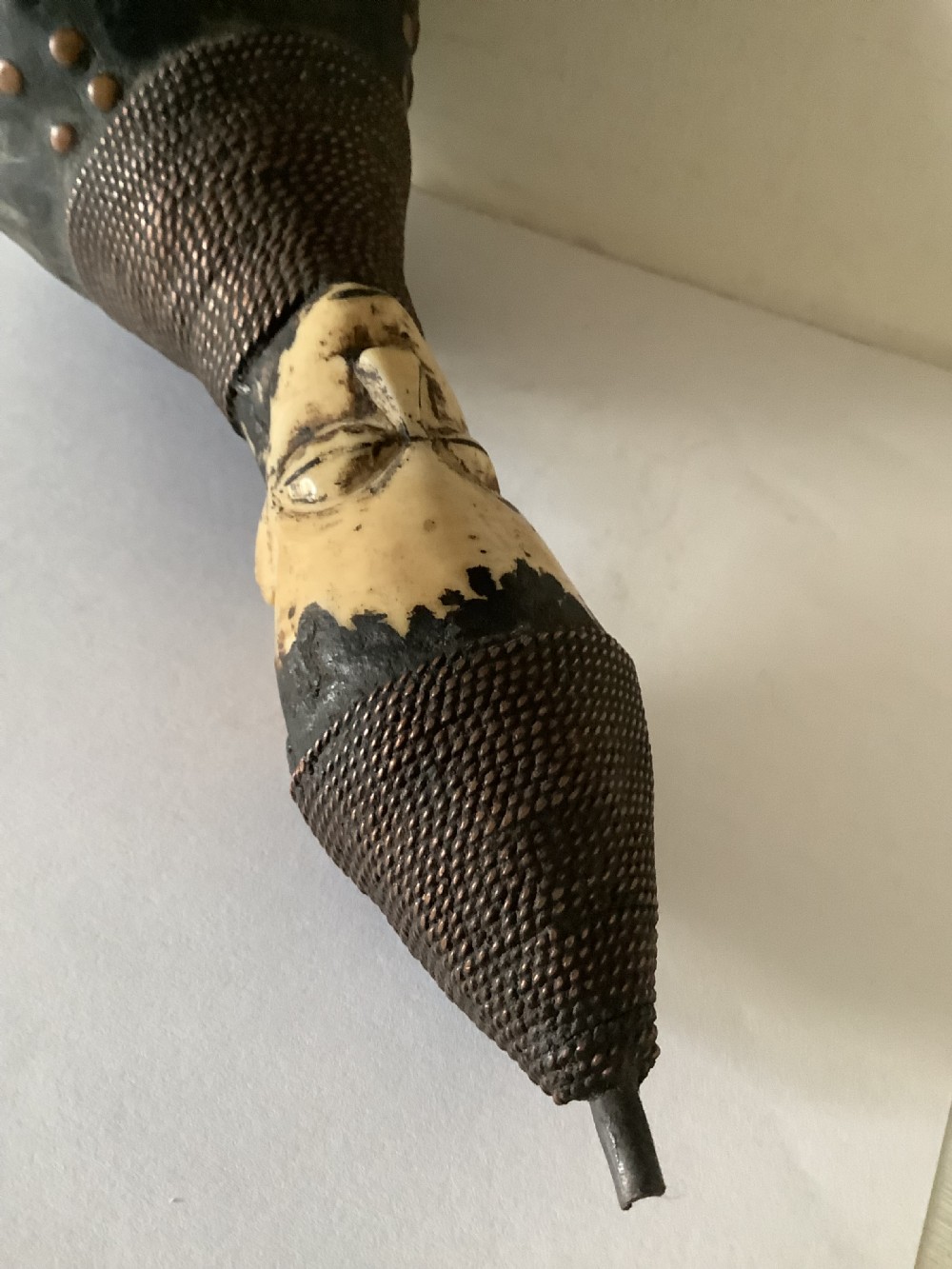A hand-carved African smoking pipe featuring a dark rounded body with metal studs, woven wire binding, and stylised carved bone faces on the bowl and stem. View VI
