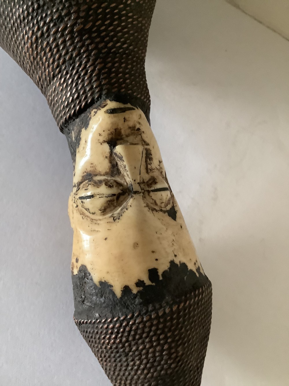A hand-carved African smoking pipe featuring a dark rounded body with metal studs, woven wire binding, and stylised carved bone faces on the bowl and stem. View VII