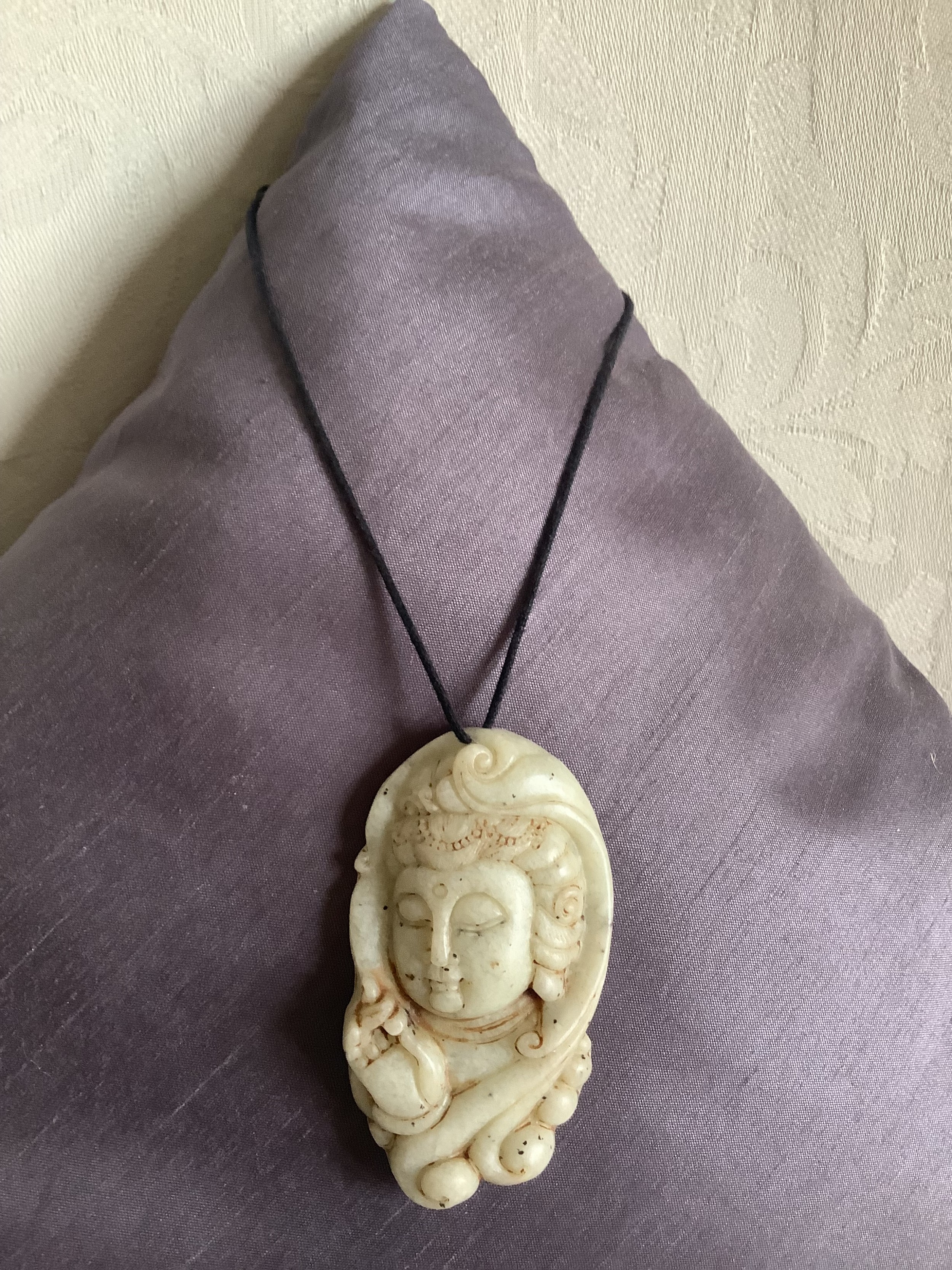 A carved pale celadon jade pendant featuring a serene deity head, suspended on a dark cord against a textured background.