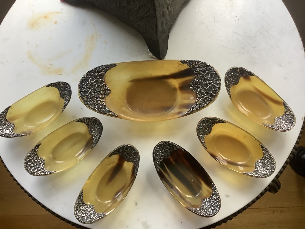 A set of seven oval amber-toned horn bowls featuring carved floral silver mounts at each end.