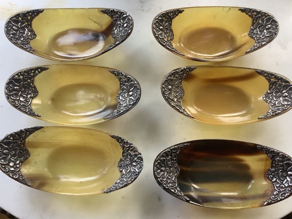 A set of seven oval amber-toned horn bowls featuring carved floral silver mounts at each end. View V