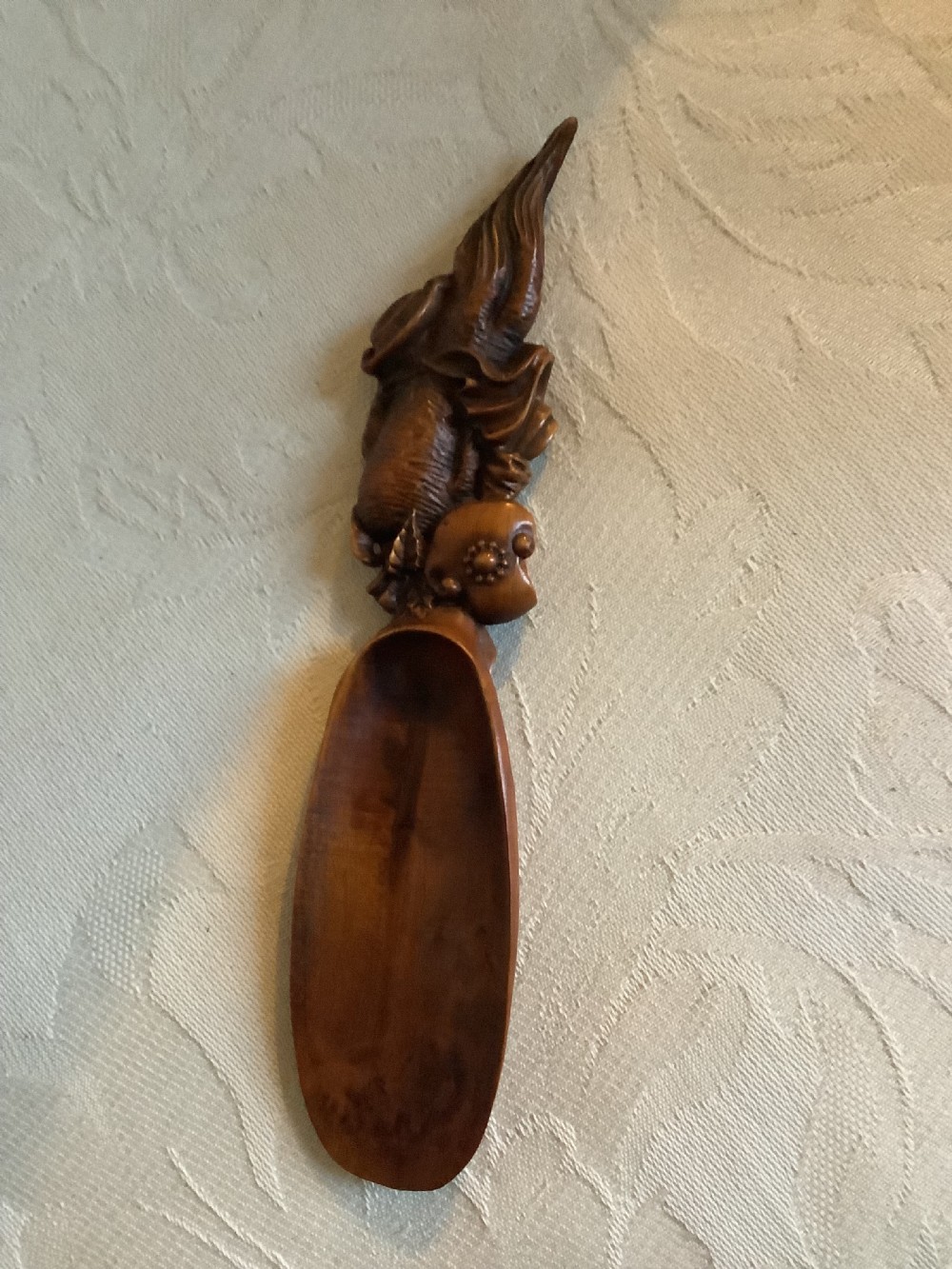 A hand-carved warm brown hardwood tea scoop featuring detailed figurative relief work along the upper handle. View II