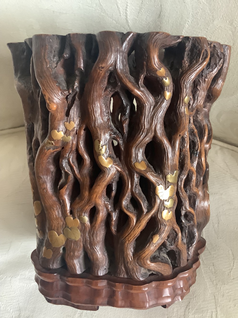 A dark brown carved hardwood brush pot featuring natural gnarled openwork root design with brass leaf inlays, resting on a carved base.