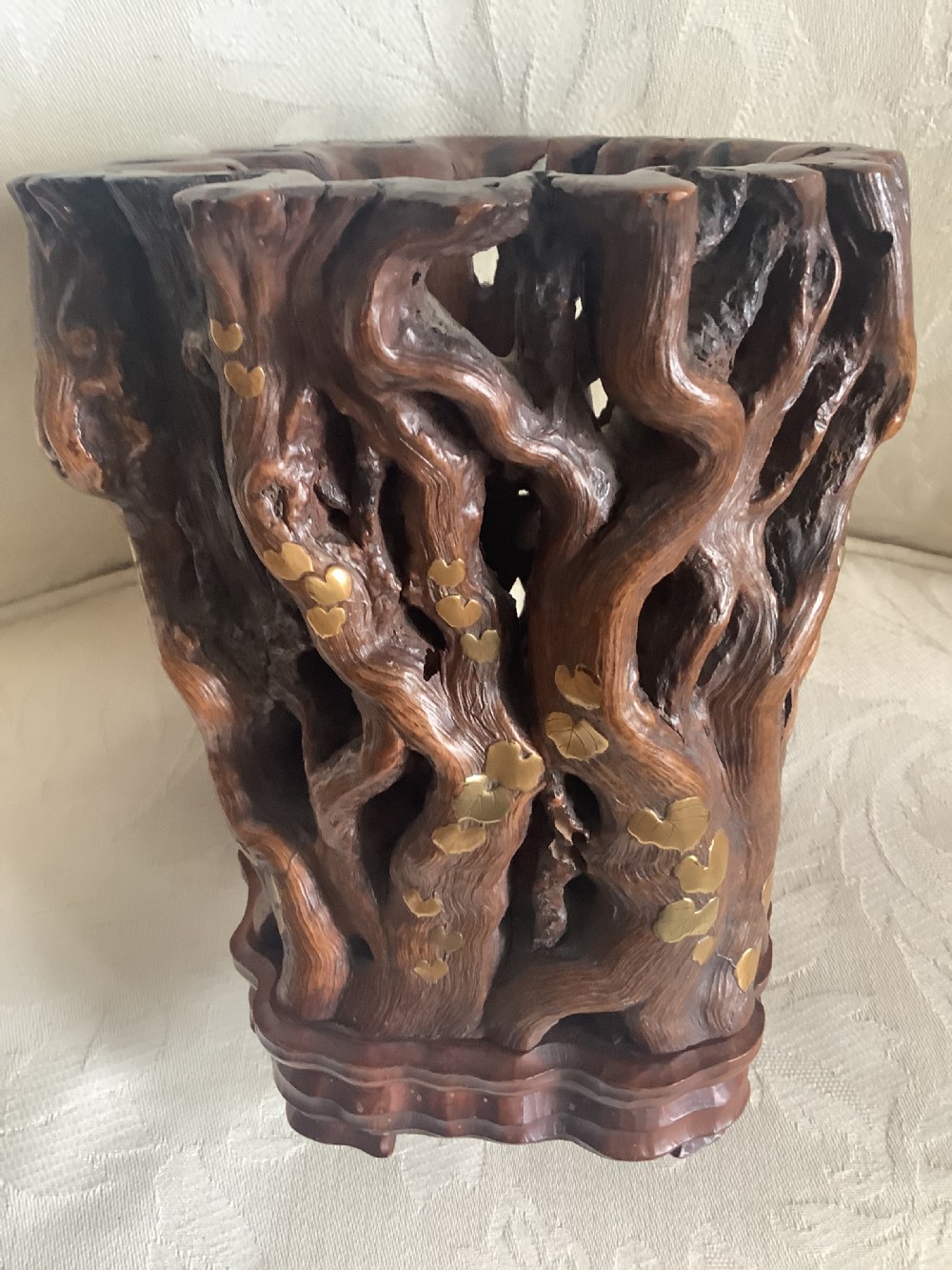 A dark brown carved hardwood brush pot featuring natural gnarled openwork root design with brass leaf inlays, resting on a carved base. View IV