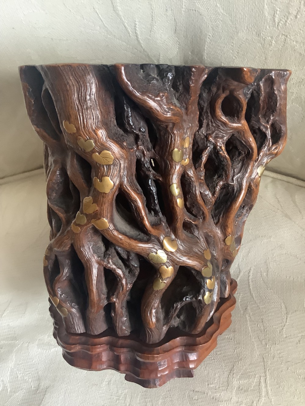 A dark brown carved hardwood brush pot featuring natural gnarled openwork root design with brass leaf inlays, resting on a carved base. View VII