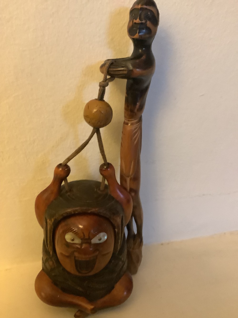 A carved wooden Japanese tobacco container featuring a wide-eyed Daruma figure connected by a cord and bead to an elongated carved wooden pipe holder. View II