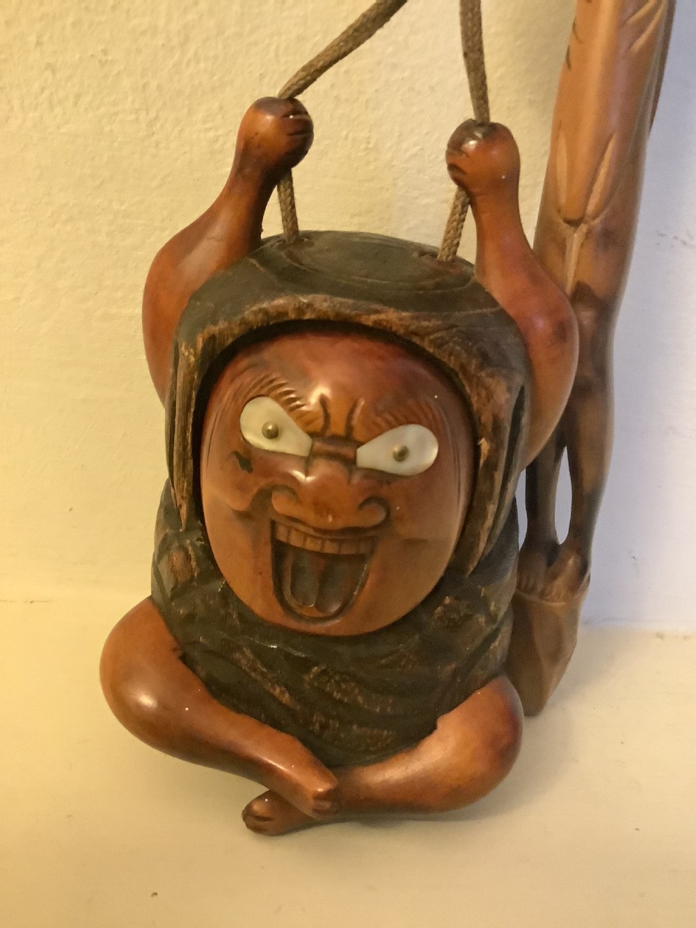 A carved wooden Japanese tobacco container featuring a wide-eyed Daruma figure connected by a cord and bead to an elongated carved wooden pipe holder. View X