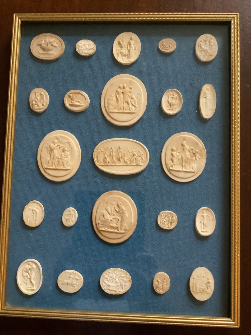 A framed arrangement of cream plaster relief medallions depicting classical figures against a blue fabric background in a gilt wooden frame.