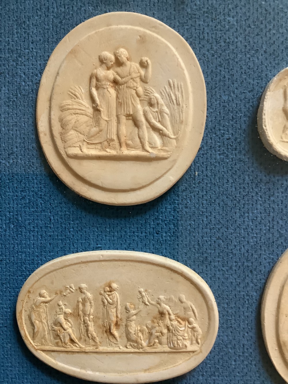 A framed arrangement of cream plaster relief medallions depicting classical figures against a blue fabric background in a gilt wooden frame. View II