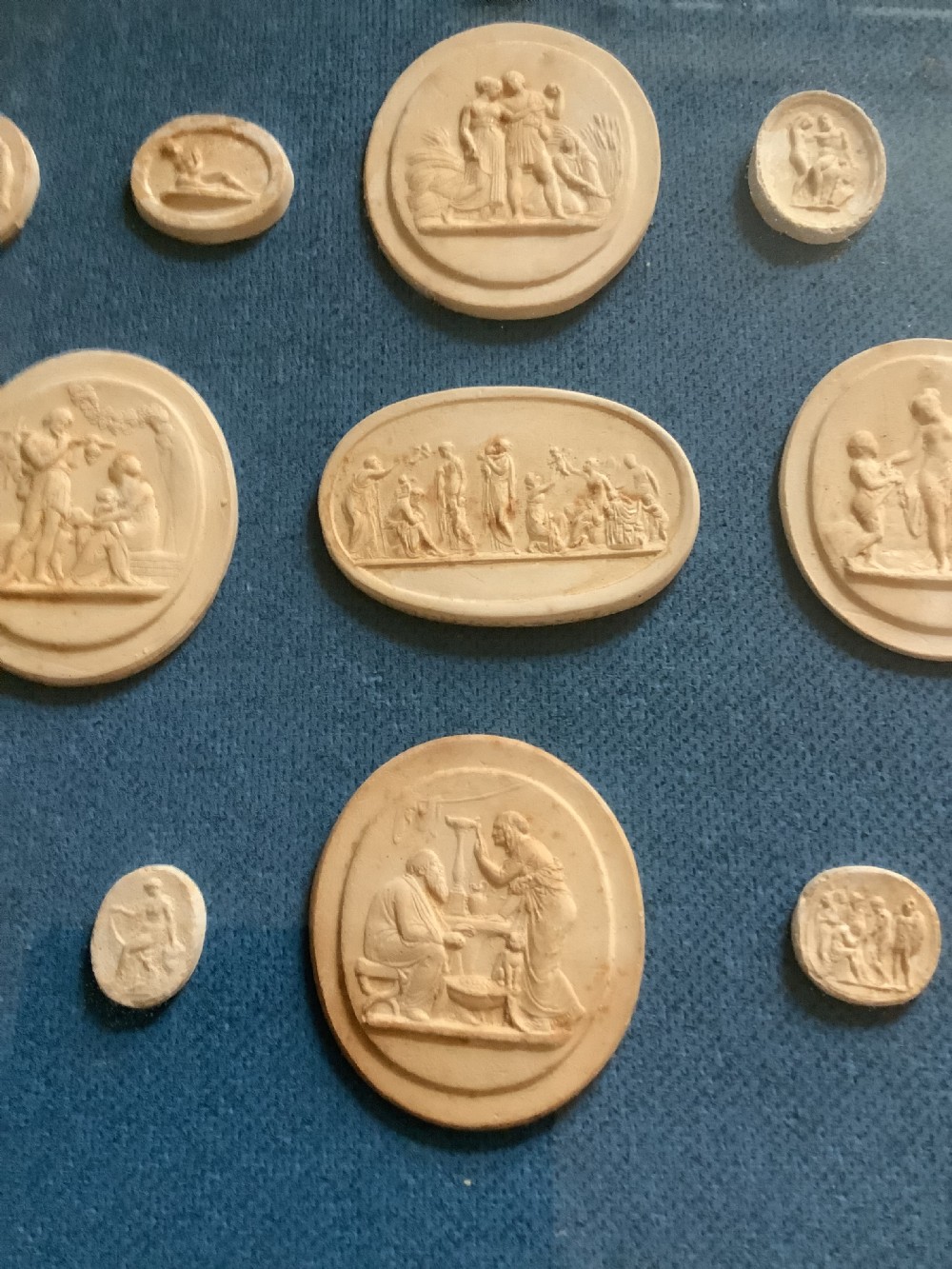 A framed arrangement of cream plaster relief medallions depicting classical figures against a blue fabric background in a gilt wooden frame. View III