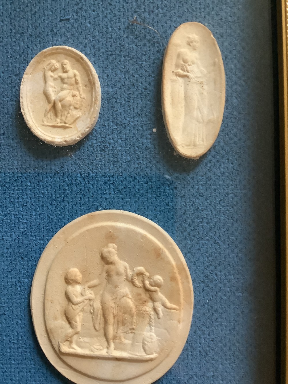 A framed arrangement of cream plaster relief medallions depicting classical figures against a blue fabric background in a gilt wooden frame. View IV
