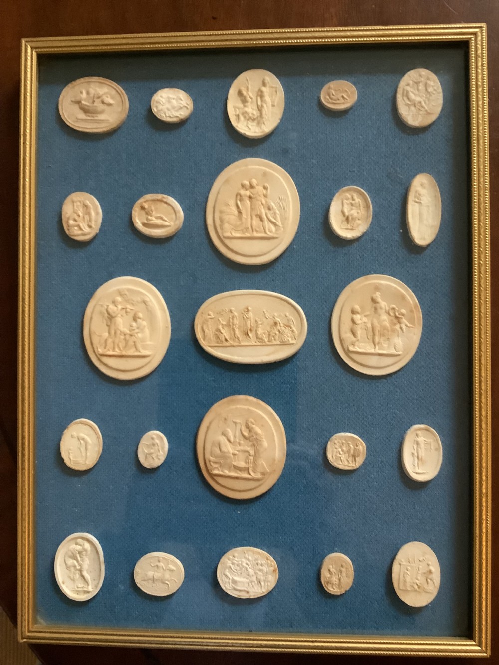 A framed arrangement of cream plaster relief medallions depicting classical figures against a blue fabric background in a gilt wooden frame. View V