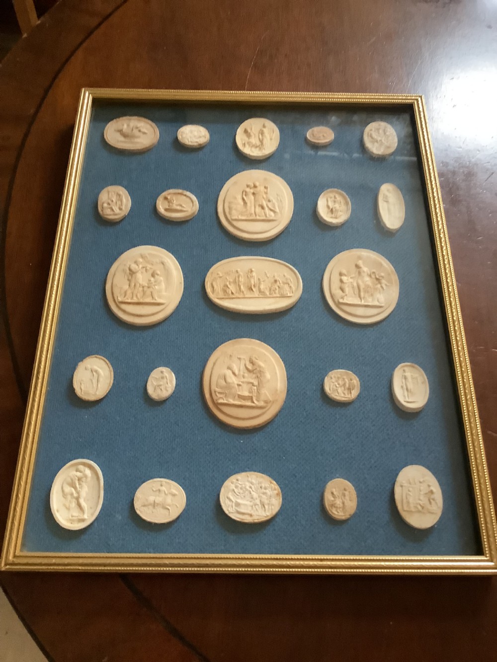 A framed arrangement of cream plaster relief medallions depicting classical figures against a blue fabric background in a gilt wooden frame. View VI