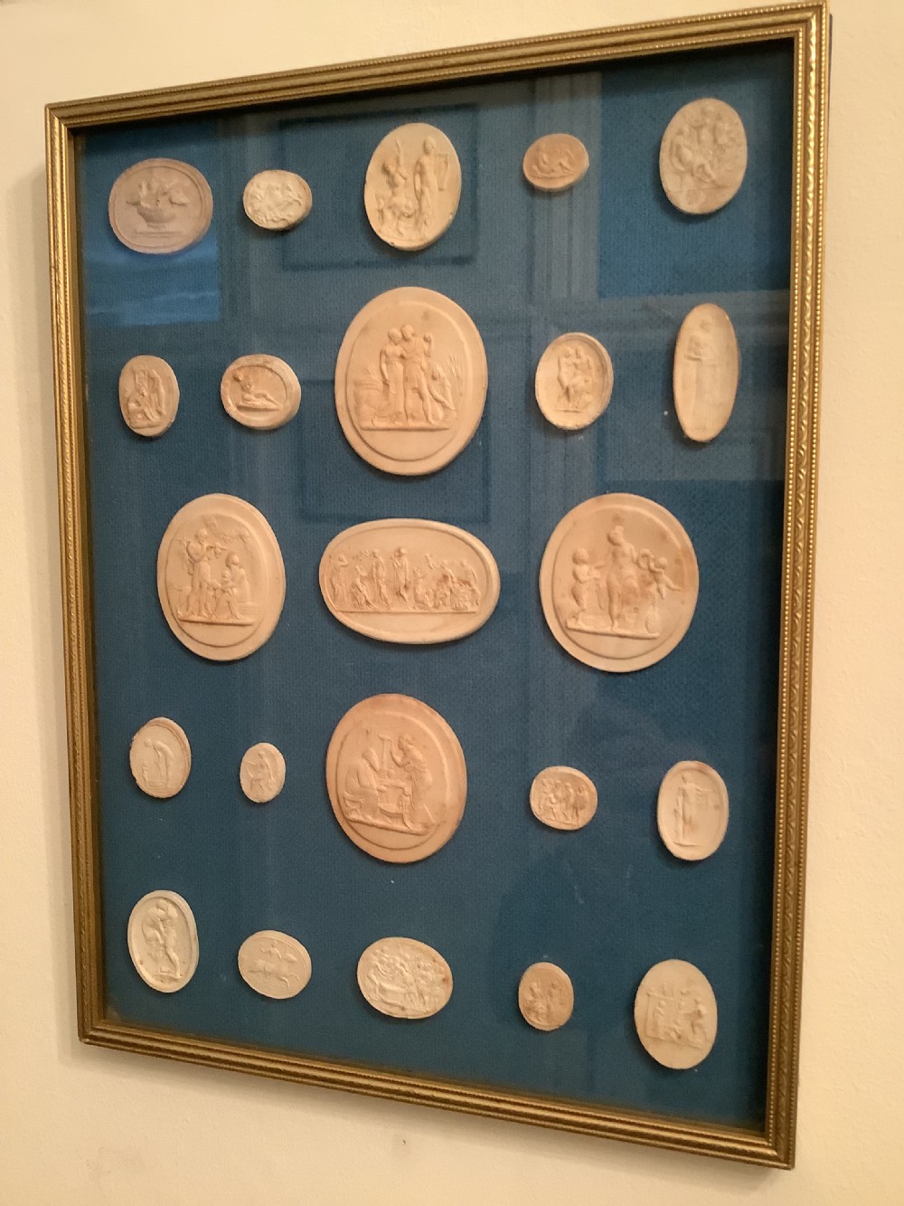 A framed arrangement of cream plaster relief medallions depicting classical figures against a blue fabric background in a gilt wooden frame. View VII