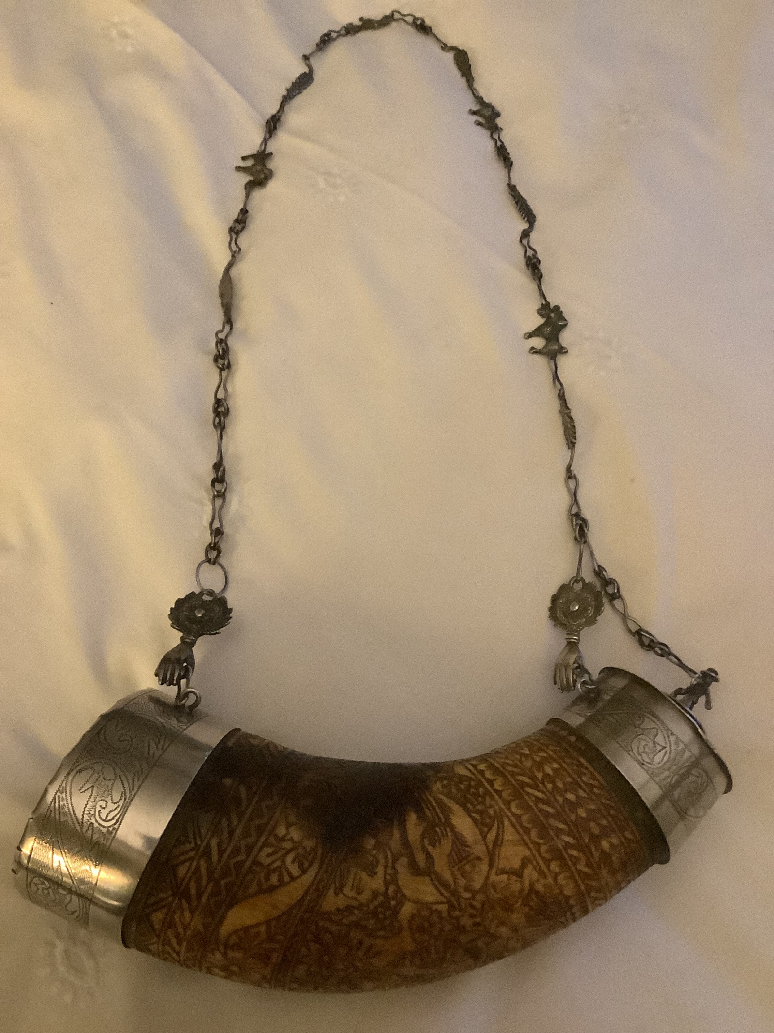 An 18th-century curved horn powder flask featuring intricate surface engravings, silver-coloured metal end caps, and an ornate chain strap.