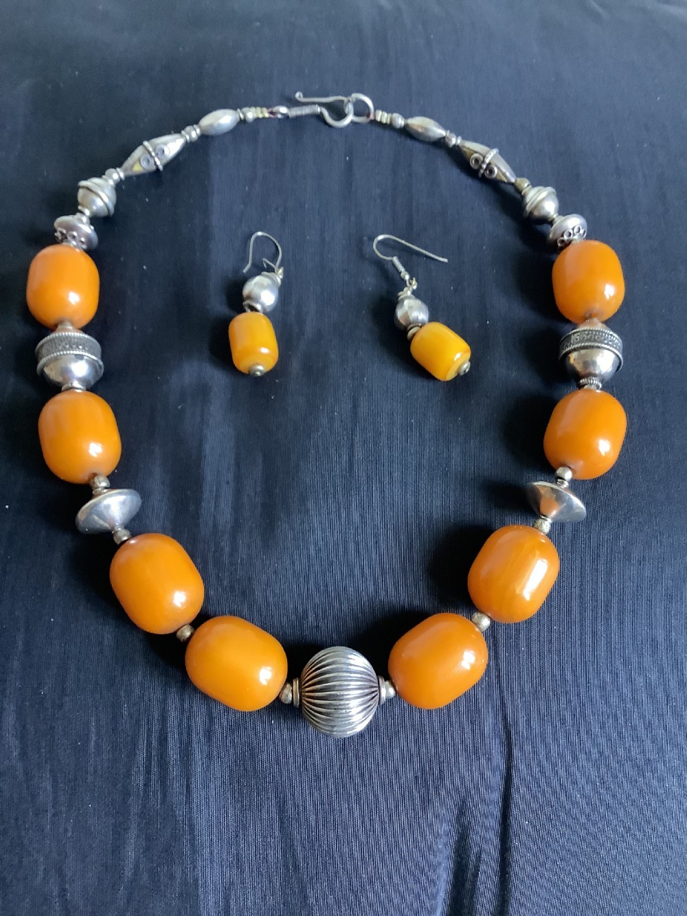 An orange amber bead and sterling silver necklace with matching drop earrings displayed flat on a dark fabric surface. View IV