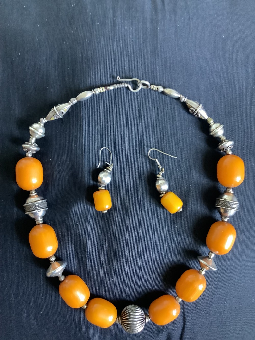 An orange amber bead and sterling silver necklace with matching drop earrings displayed flat on a dark fabric surface. View V